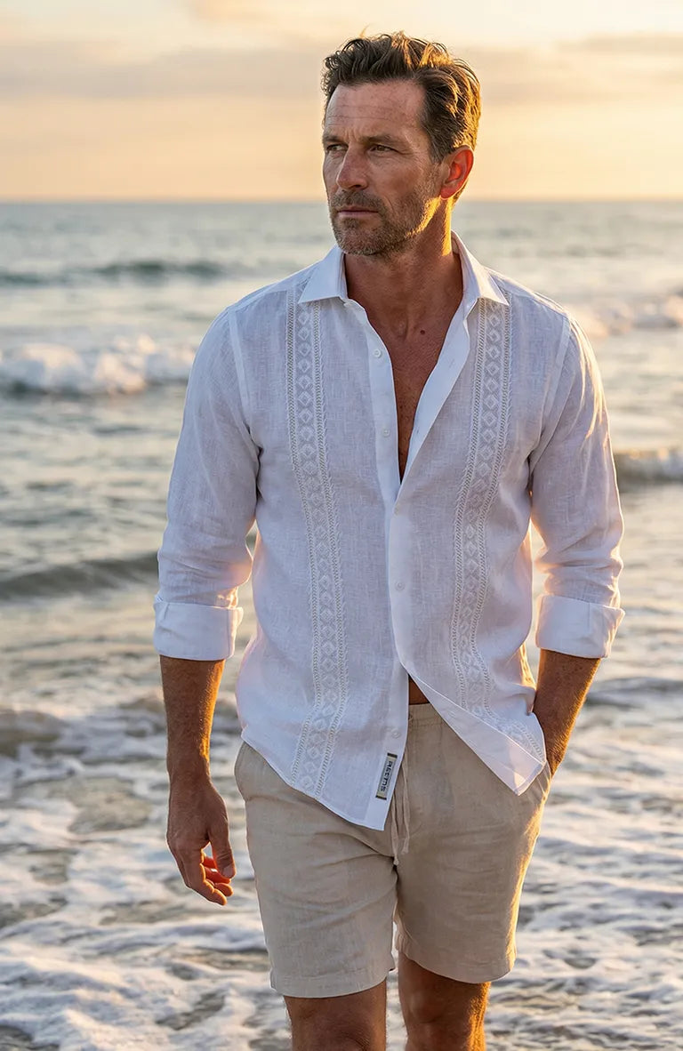 Man wearing a white Guayabera Shirt and beige shorts standing on a beach at sunset.